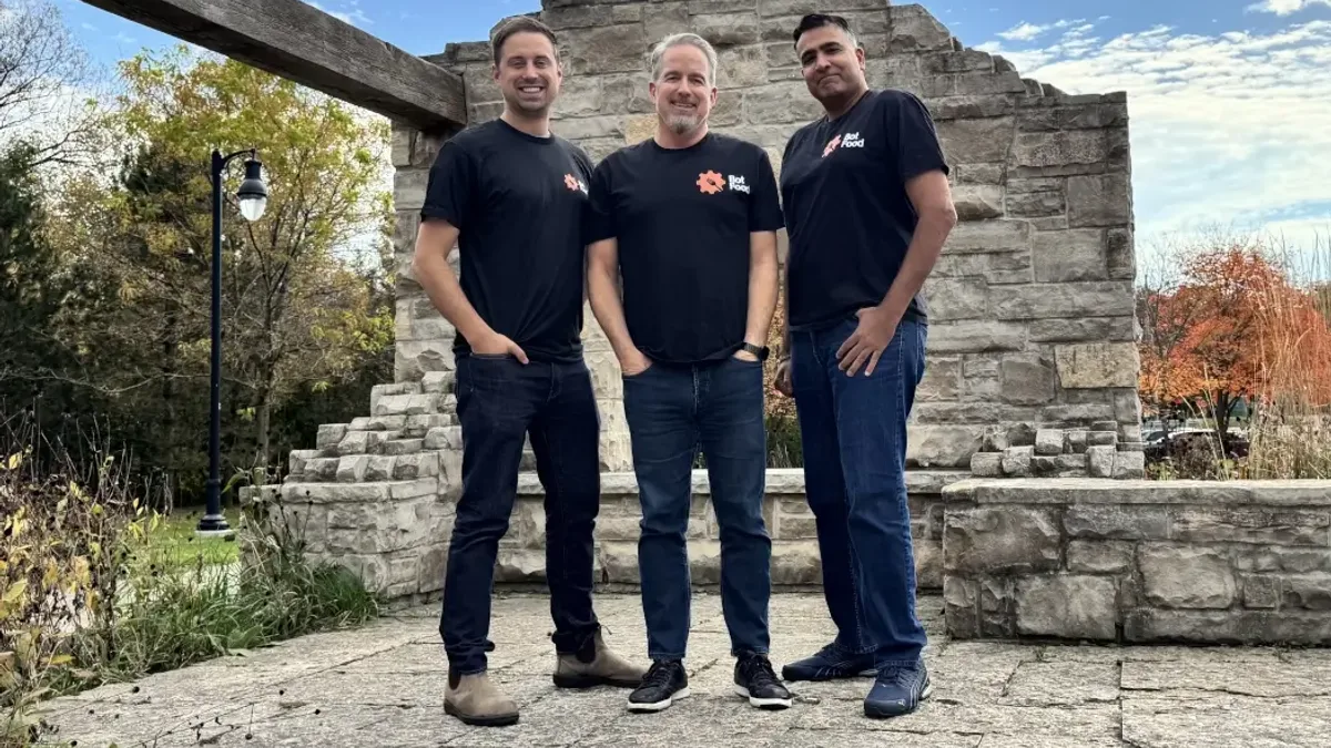 Three members of the Bot Food team in black Bot Food T-shirts, standing in front of a stone wall in a park.